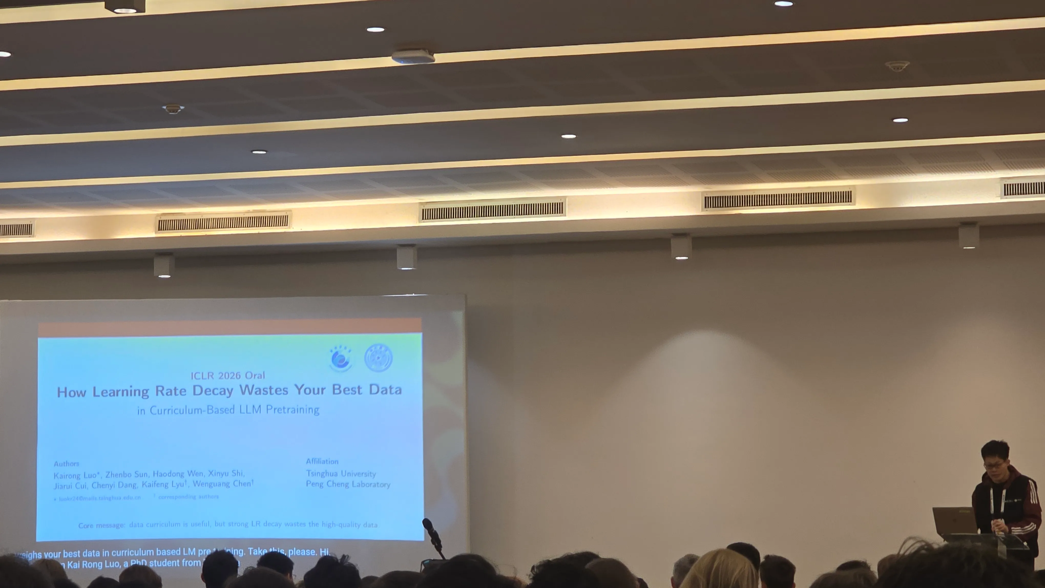 Conference stage. Projected title slide reads 'ICLR 2026 Oral, How Learning Rate Decay Wastes Your Best Data in Curriculum-Based LLM Pretraining' with author list Kairong Luo, Zhenbo Sun, Haodong Wen, Xinyu Shi, Jiarui Cui, Chenyi Dang, Kaifeng Lyu, Wenguang Chen, affiliated with Tsinghua University and Peng Cheng Laboratory. A speaker in a red jacket stands at a podium to the right of the screen.
